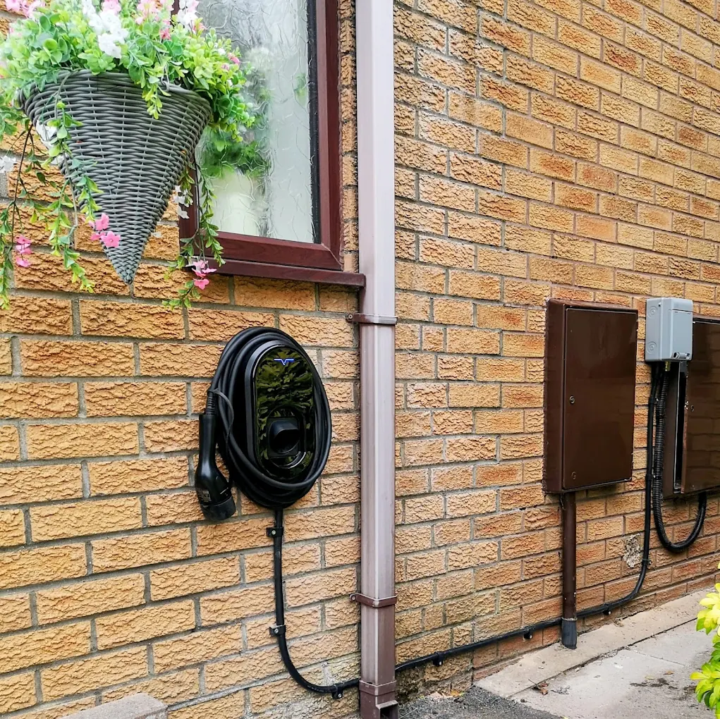 Black EV charger with coiled cable installed on a brick wall beside a hanging basket